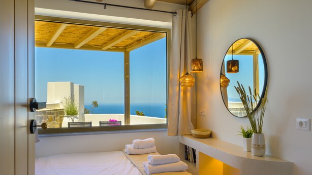 Habitación de hotel con un ventanal grande, una cama con toallas dobladas con esmero, un espejo redondo y una pequeña planta en maceta.