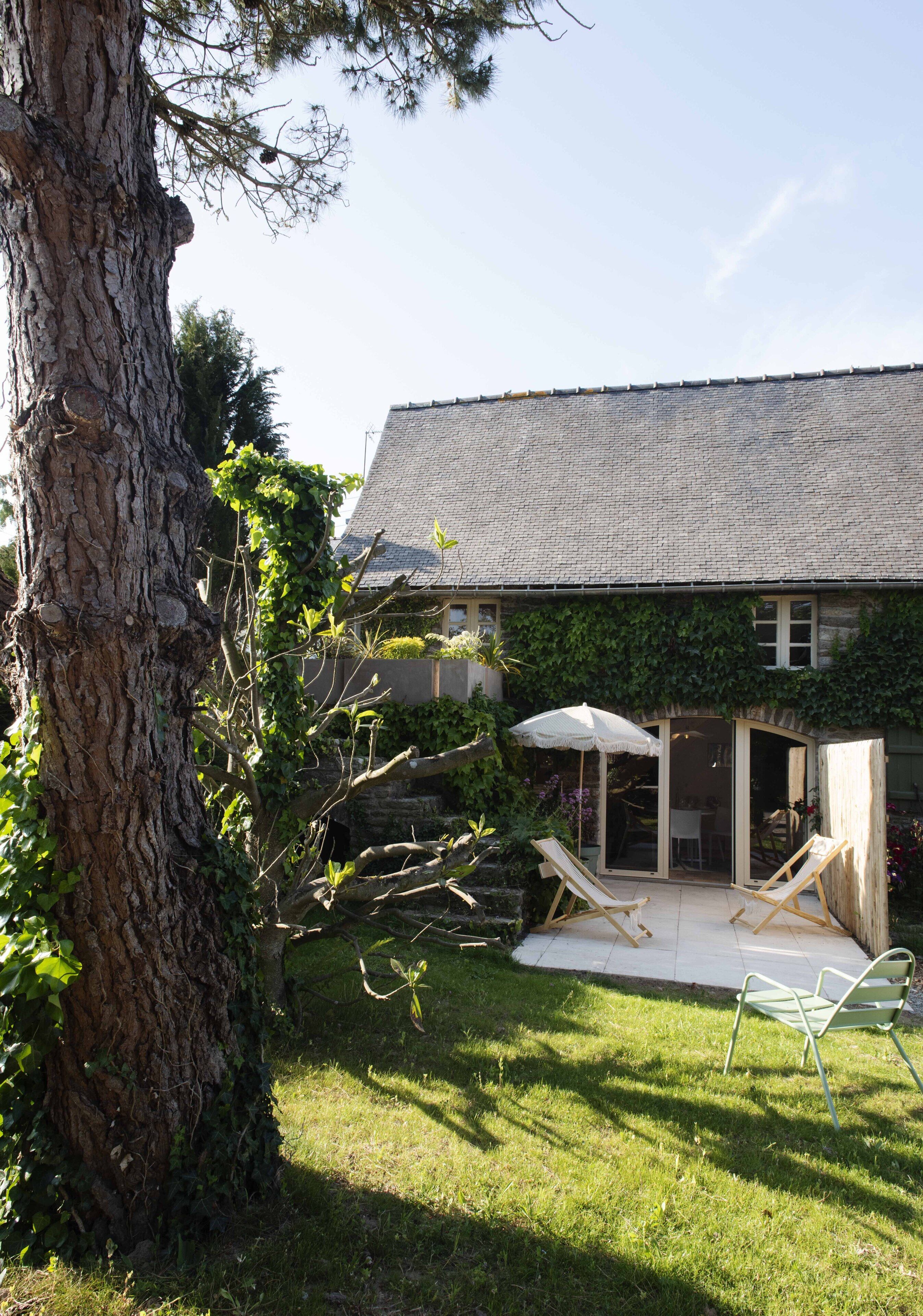 A house with a sloped roof, a patio with two lounge chairs and a table, and a large tree in the foreground.