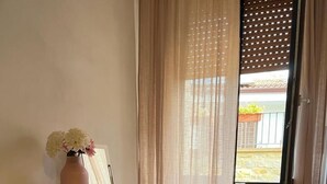 A hotel room with a bed, a desk with a computer, a chair, a small heater, and a window with curtains.