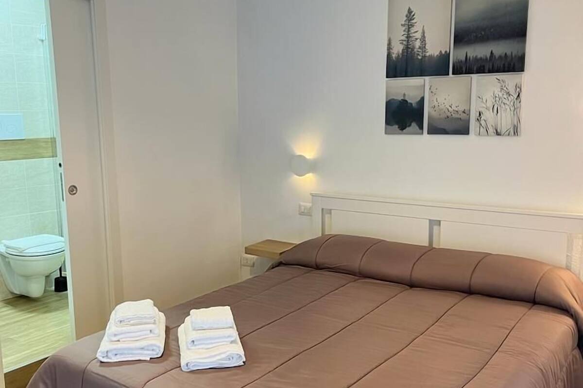 A neatly made bed with a brown cover, a bedside table, and a wall with four landscape paintings.
