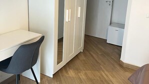 A room with a white wardrobe, a desk with a chair, and a bed.