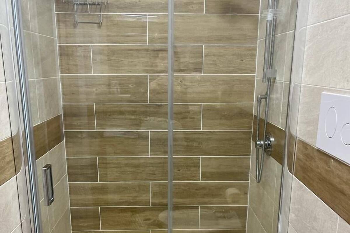 A glass-enclosed shower stall with a beige tiled floor and a white toilet.