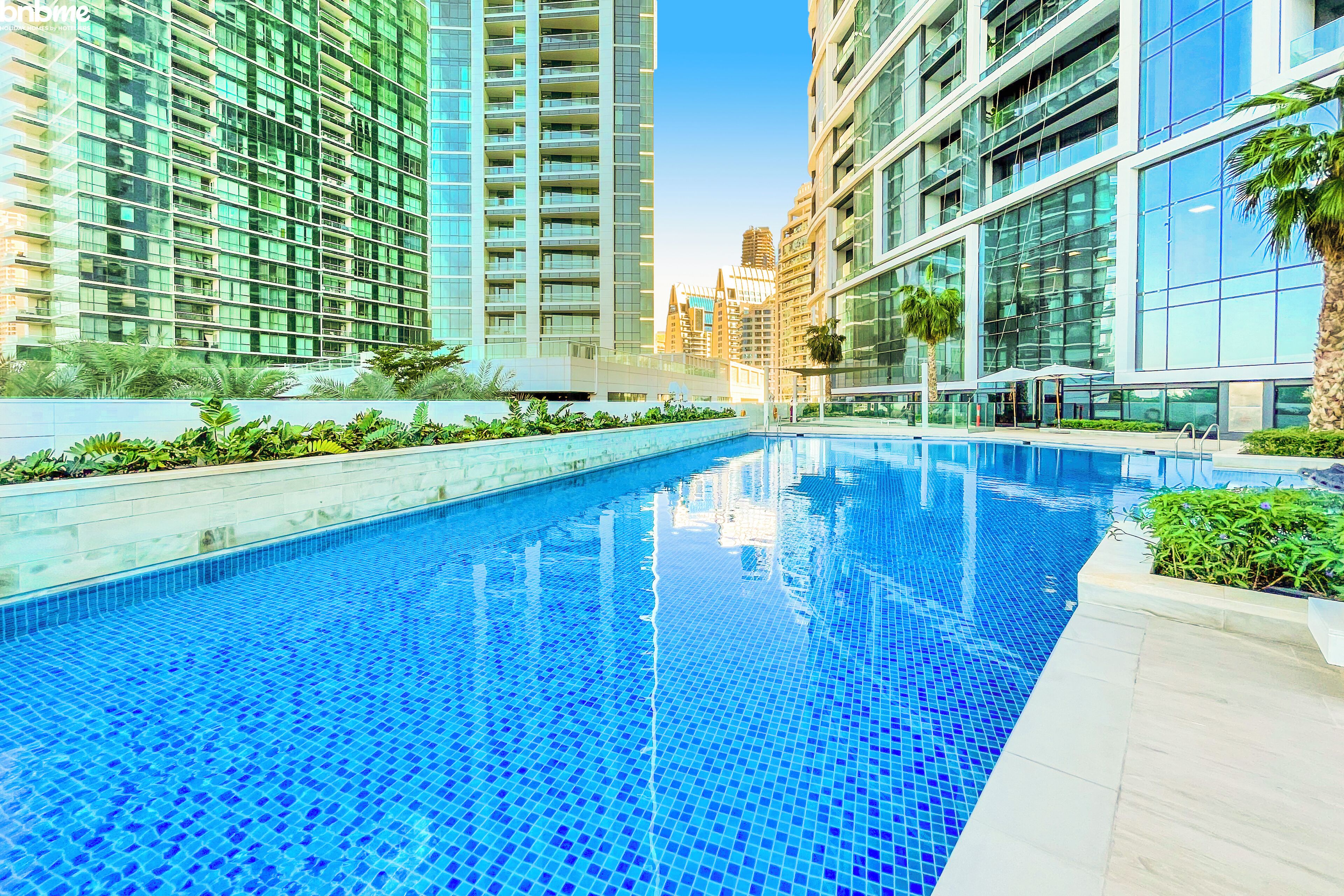 A swimming pool with clear blue tiles, surrounded by modern buildings with glass facades and a well-maintained garden area.