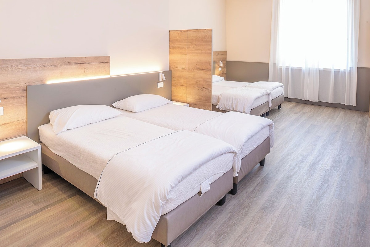 A hotel room with two beds, a wooden headboard, a nightstand, and a window with sheer curtains.