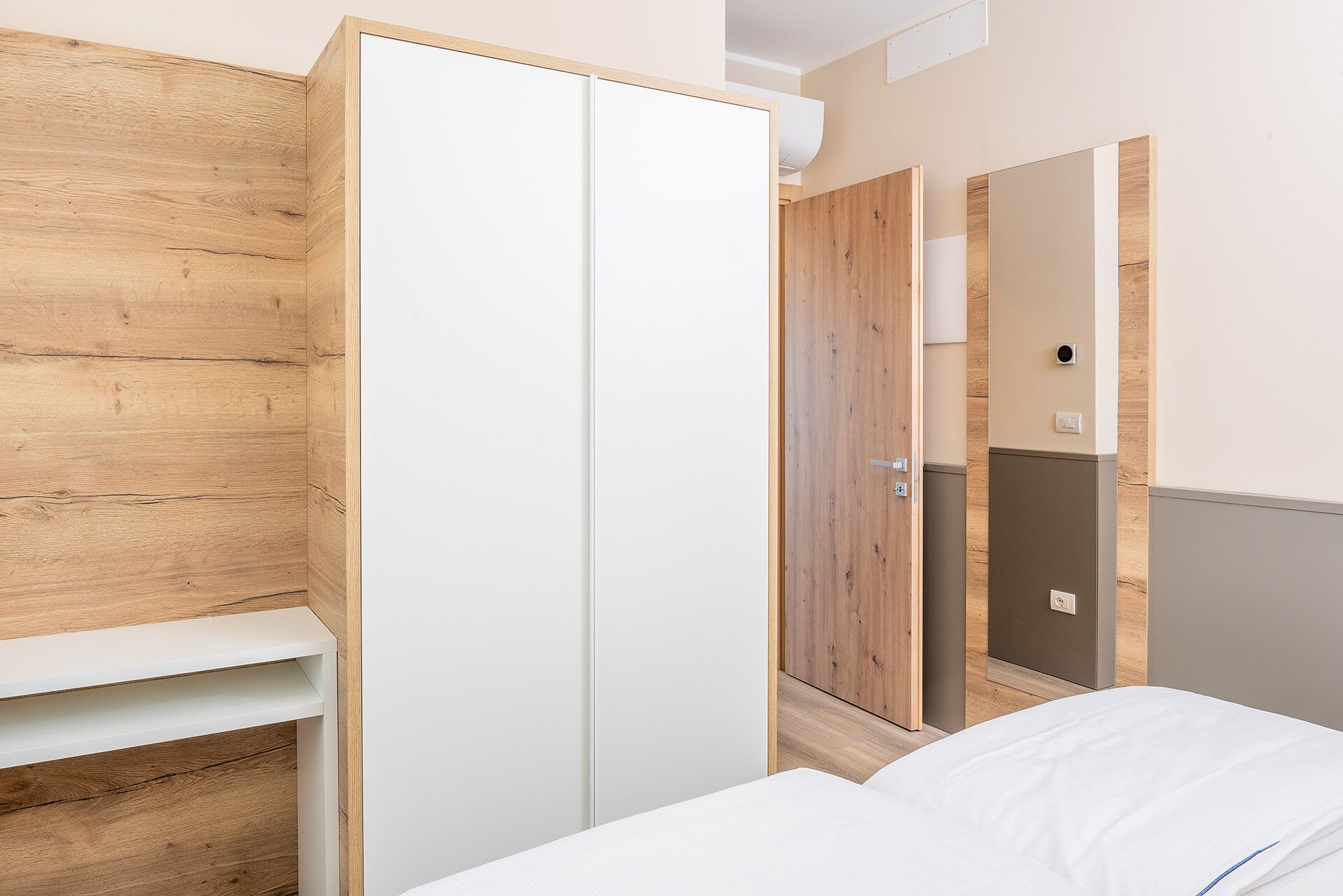 A bedroom with a wooden wardrobe, a white bed, and a mirror reflecting the room.