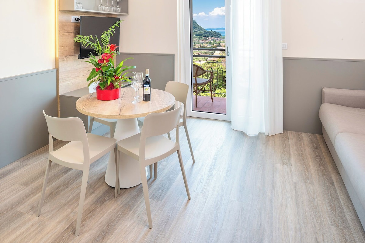 A modern apartment with a dining area featuring a round table, white chairs, and a red flower vase. A balcony with a view of a hillside town is visible through a glass door.