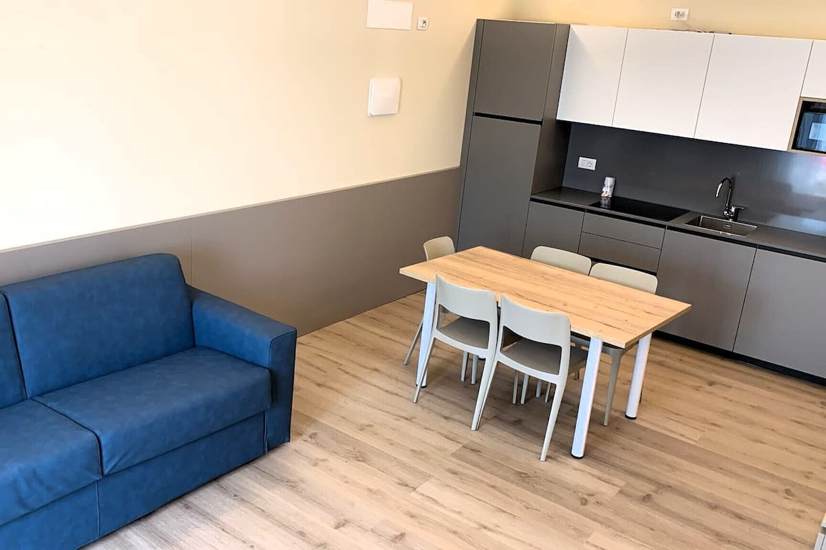 A compact living space with a blue sofa, a wooden dining table with four chairs, a kitchenette, and a light-colored floor.
