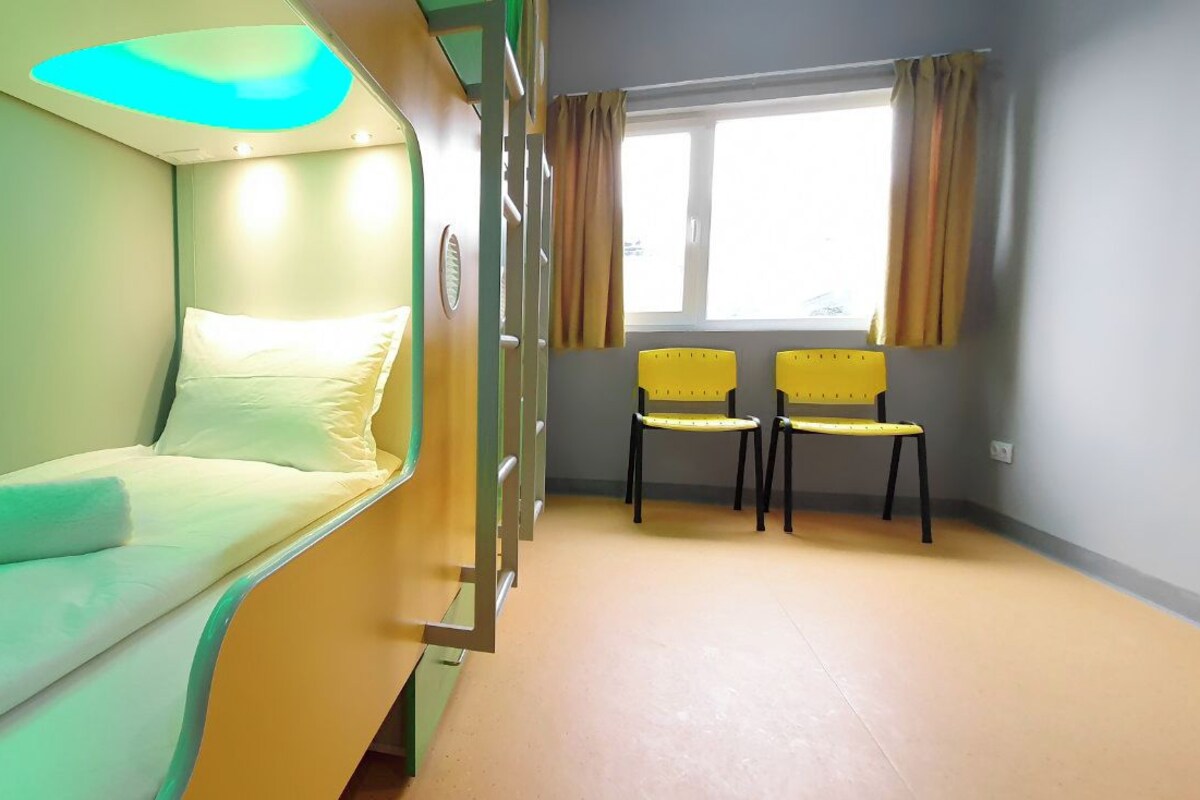 Quarto com beliches, duas cadeiras, uma janela com cortinas e um lustre.