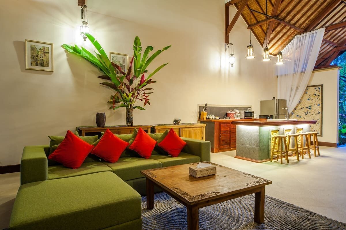 A modern living room with a green sofa, red cushions, a wooden coffee table, and a kitchen area with bar stools.
