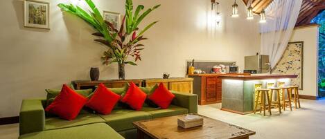 A modern living room with a green sofa, red cushions, a wooden coffee table, and a kitchen area with bar stools.