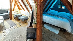 Ein Schlafzimmer im Loft-Stil mit freiliegenden Holzbalken, einem Bett, einem Fernseher und einer kleinen Sitzecke mit Kissen.