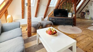 Ein Loft-Wohnzimmer mit freiliegenden Holzbalken, einem weißen Sofa, einem Couchtisch mit Obst und einem Fernseher.
