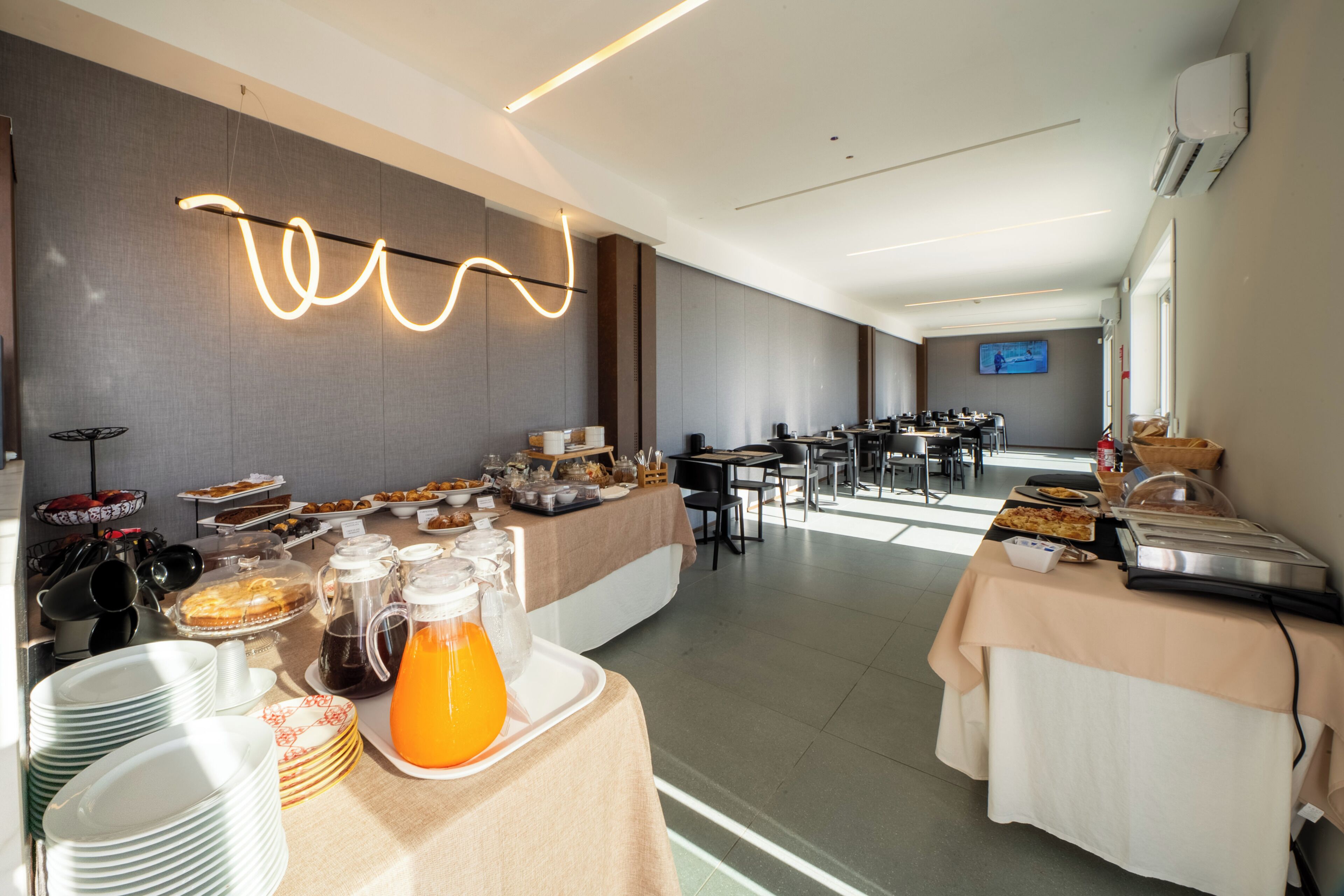 A buffet setup with various food items, a beverage dispenser, and a television screen in the background.