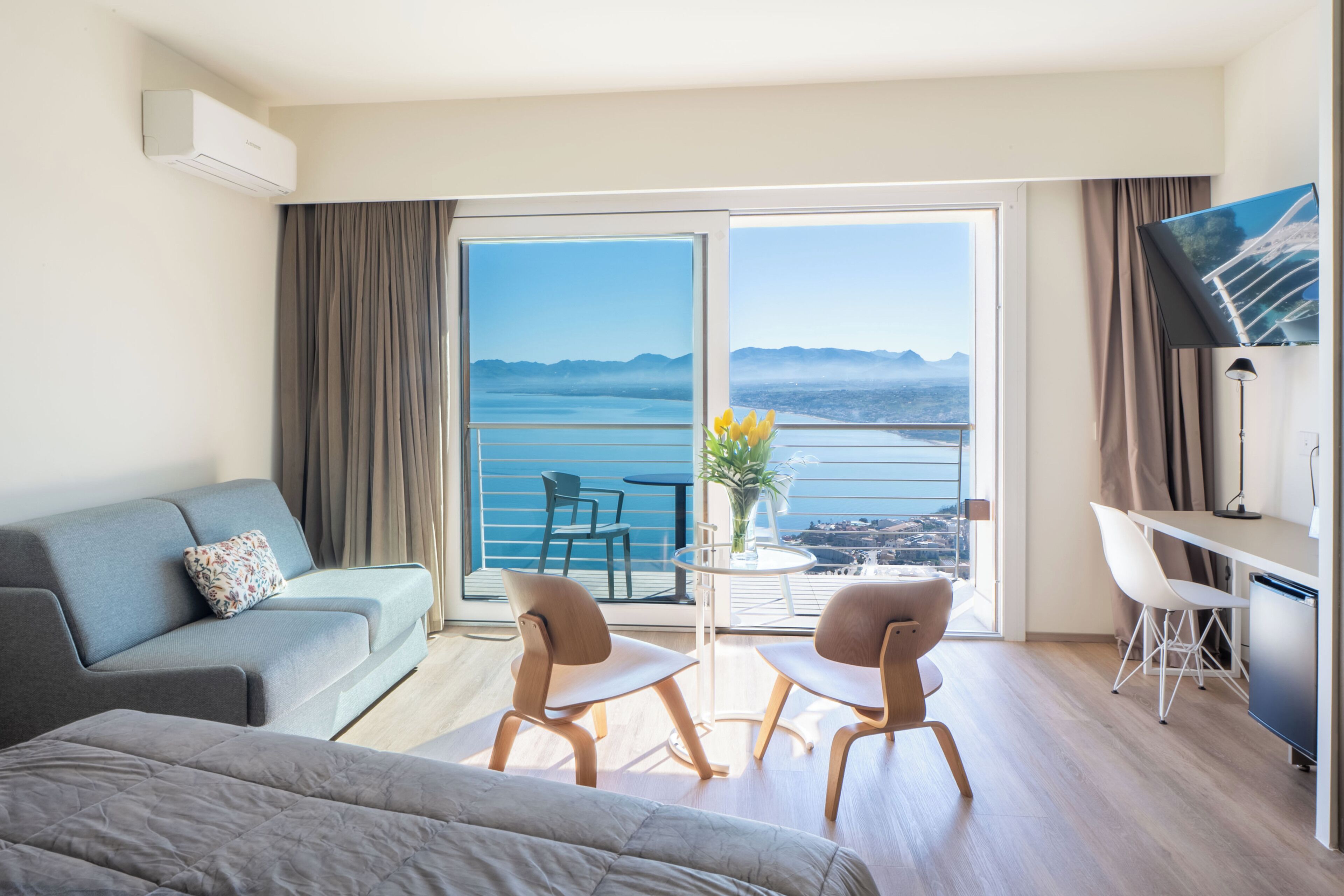 Ein modernes Hotelzimmer mit Balkon, einer Couch, zwei Sesseln, einem kleinen Tisch mit einer Vase und einem Fernseher.