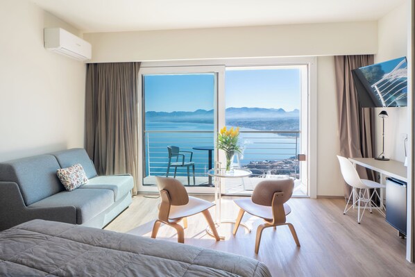 A modern hotel room with a balcony, a sofa, two chairs, a small table with a vase, and a TV.