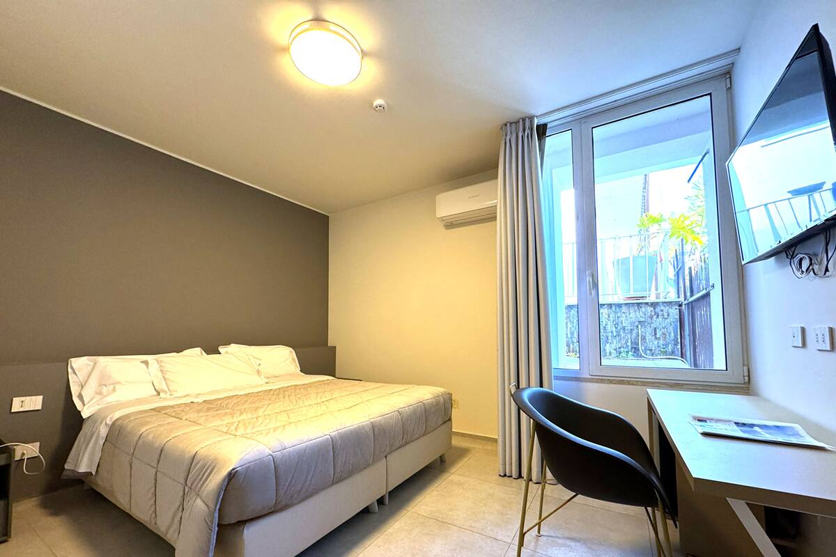 Ett hotellrum med en säng, ett skrivbord, en stol, ett fönster med gardiner och en luftkonditioneringsanläggning.
