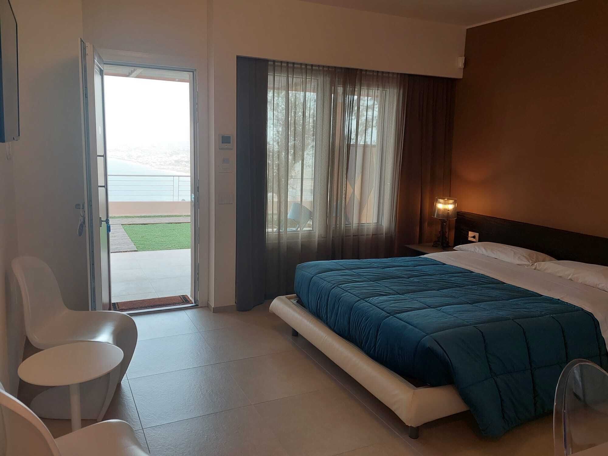 A hotel room with a bed, a chair, a table, a window with curtains, and a view of the outdoors.