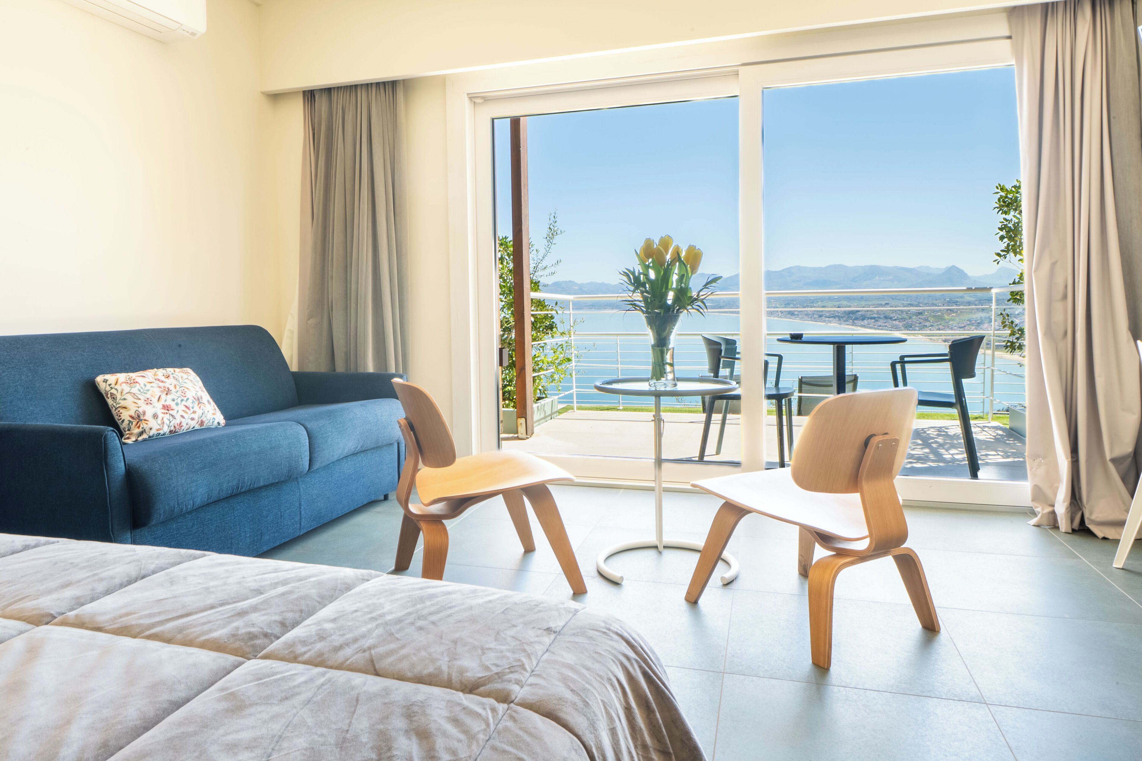 A hotel room with a bed, a blue sofa, two wooden chairs, a small table, and a balcony with a view of the sea and mountains.