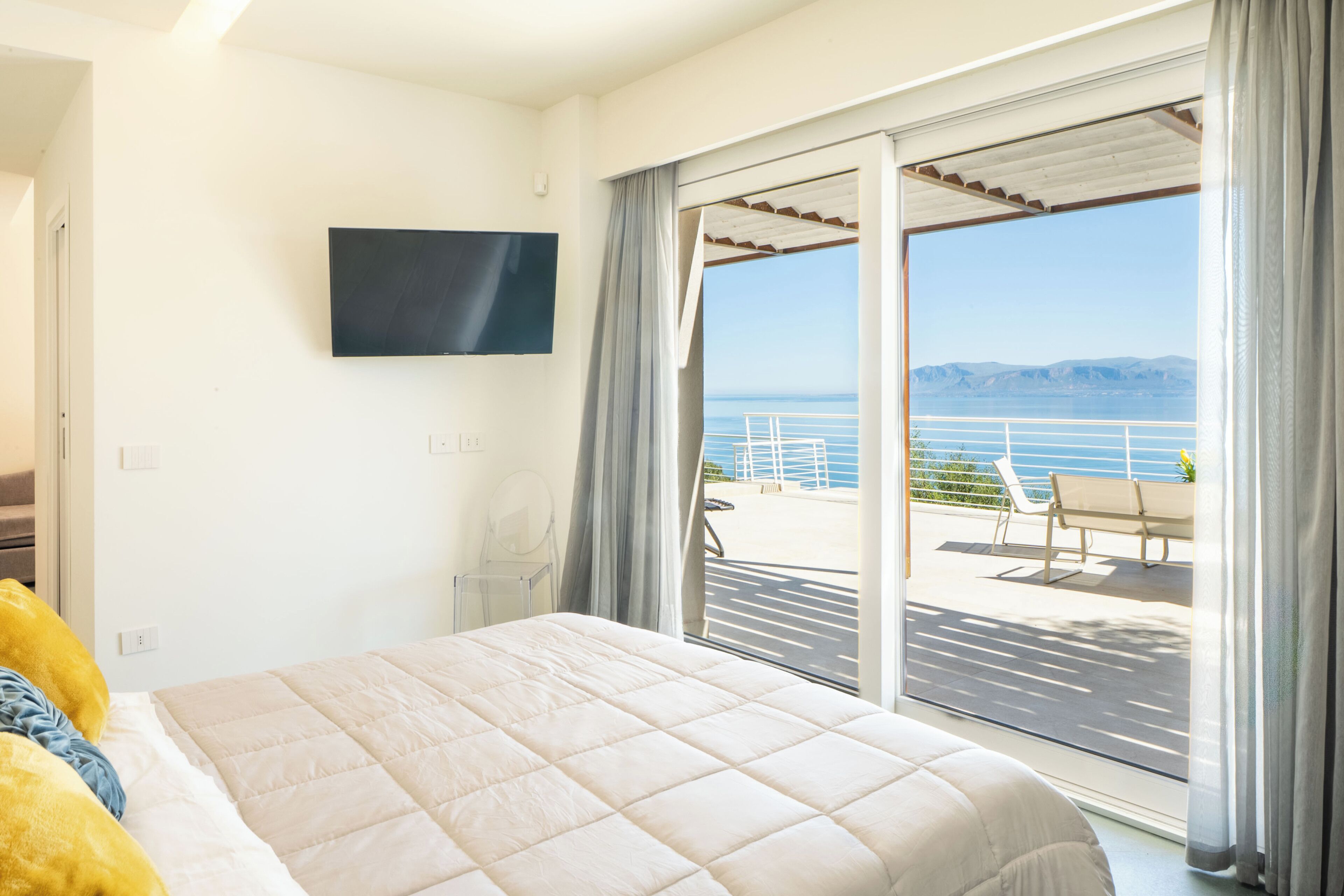 A bedroom with a bed, a TV mounted on the wall, and a balcony with a view of the sea.