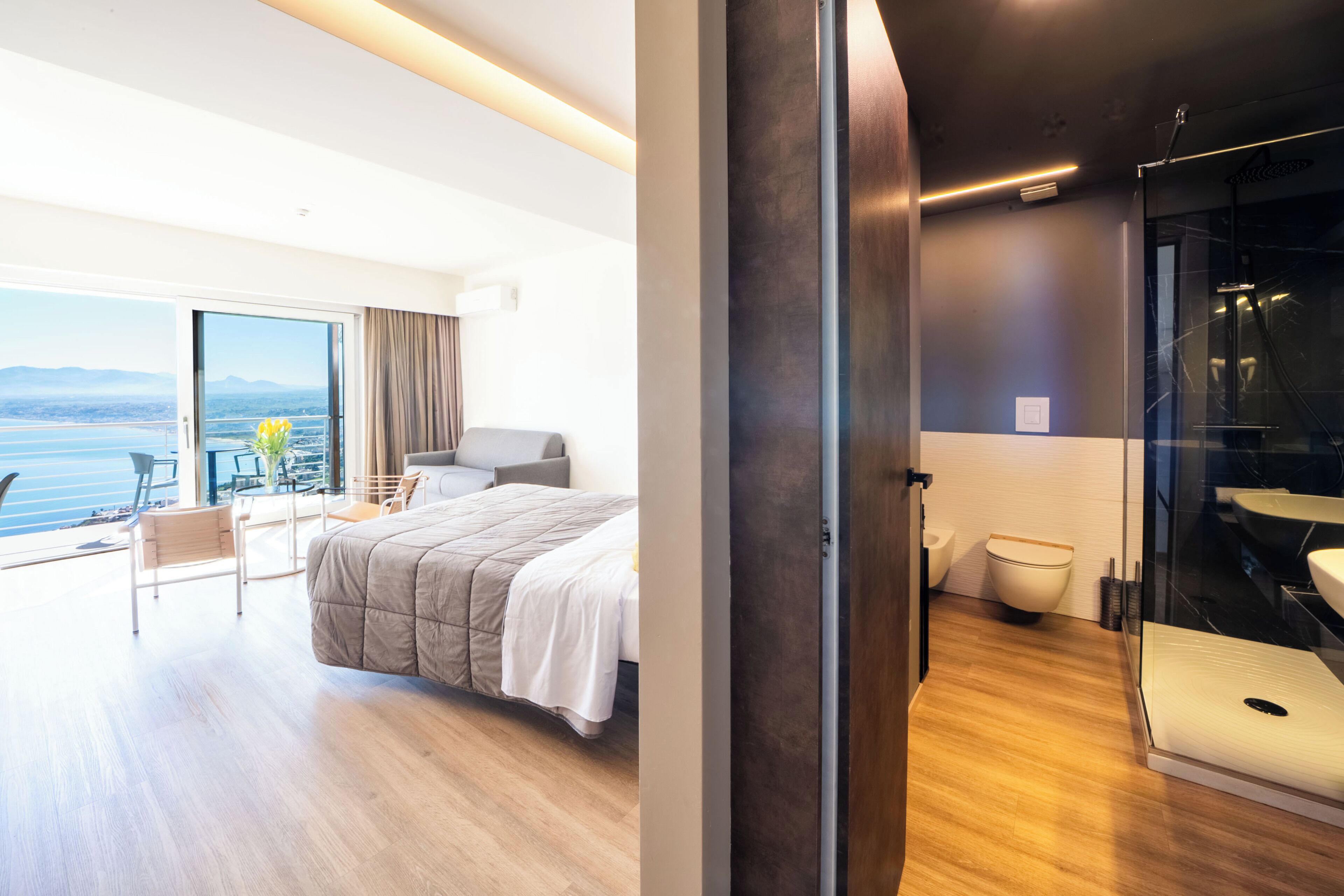 Una habitación de hotel moderna con una cama grande, una zona de estar con vistas y un baño separado con ducha de vidrio.