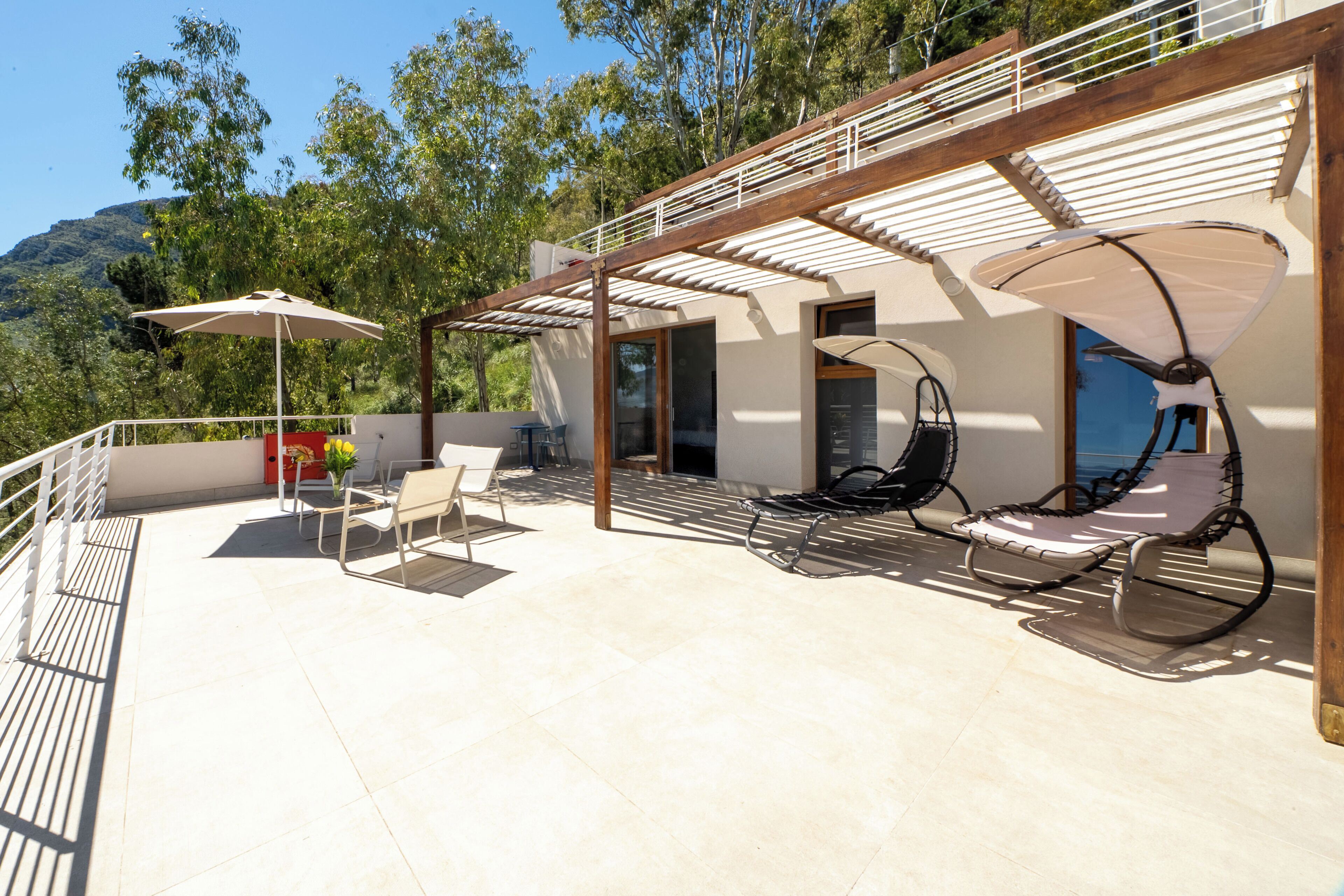 A covered outdoor seating area with chairs and umbrellas, a wooden pergola, and a view of trees and hills.