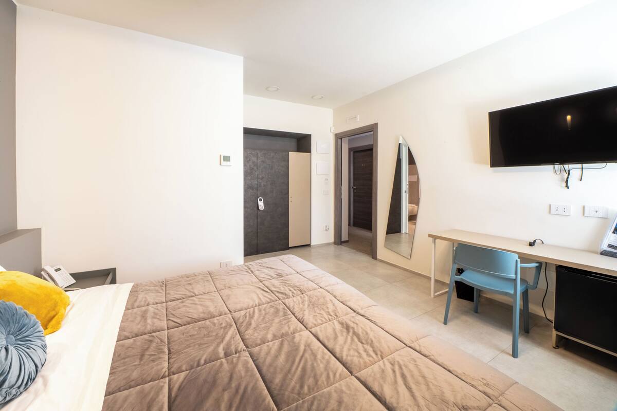 Ett modernt hotellrum med en stor säng, ett skrivbord, en stol, en platt-TV och en dörr som leder till ett annat rum.