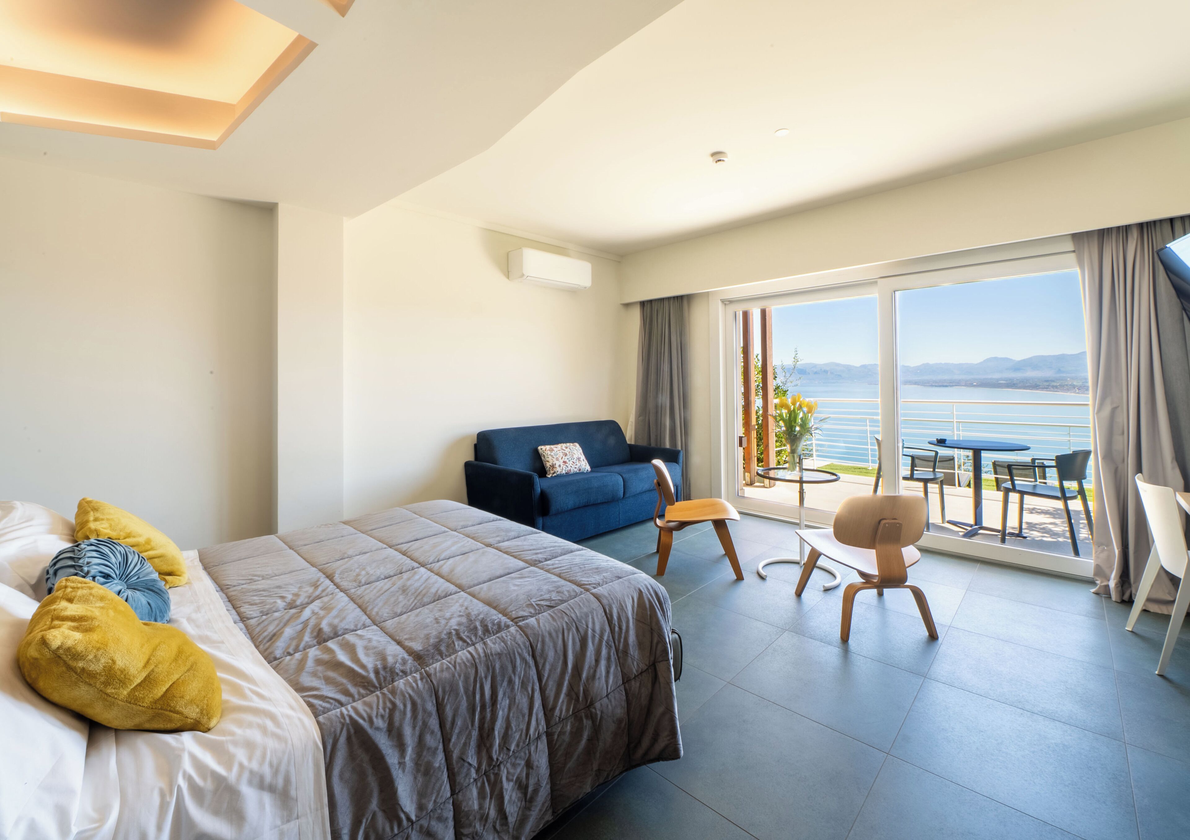 Habitación de hotel con cama, sofá, balcón con mesa y sillas, y vista al mar.