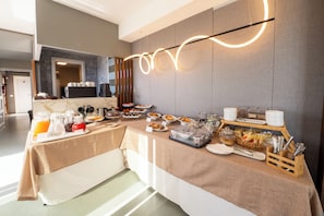 A breakfast buffet with various food items and beverages, a modern light fixture, and a kitchen area in the background.