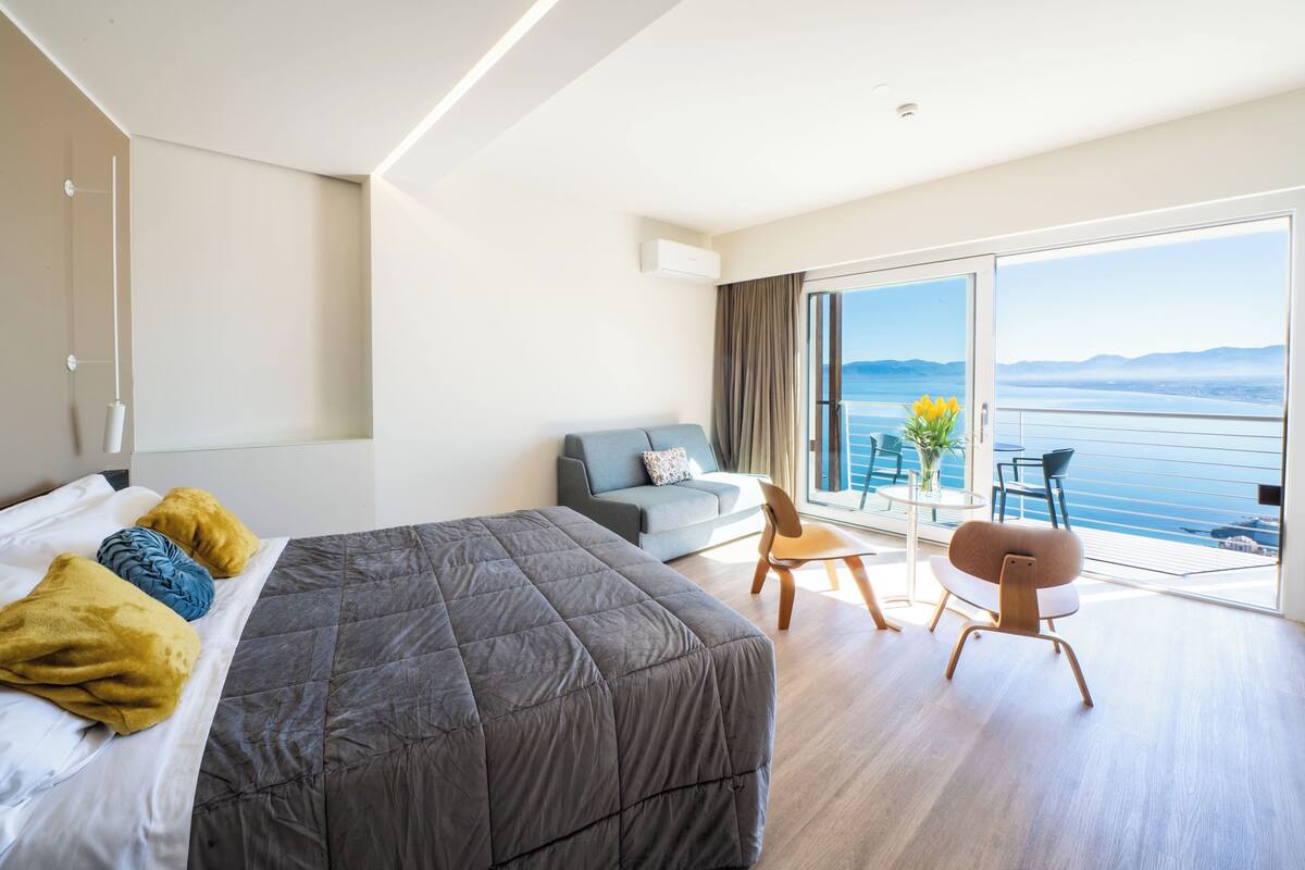Ett modernt hotellrum med en stor säng, en soffa, ett matbord och en balkong med utsikt över havet.