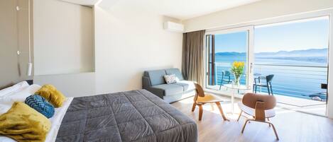 A modern hotel room with a large bed, a sofa, a dining table, and a balcony with a view of the sea.