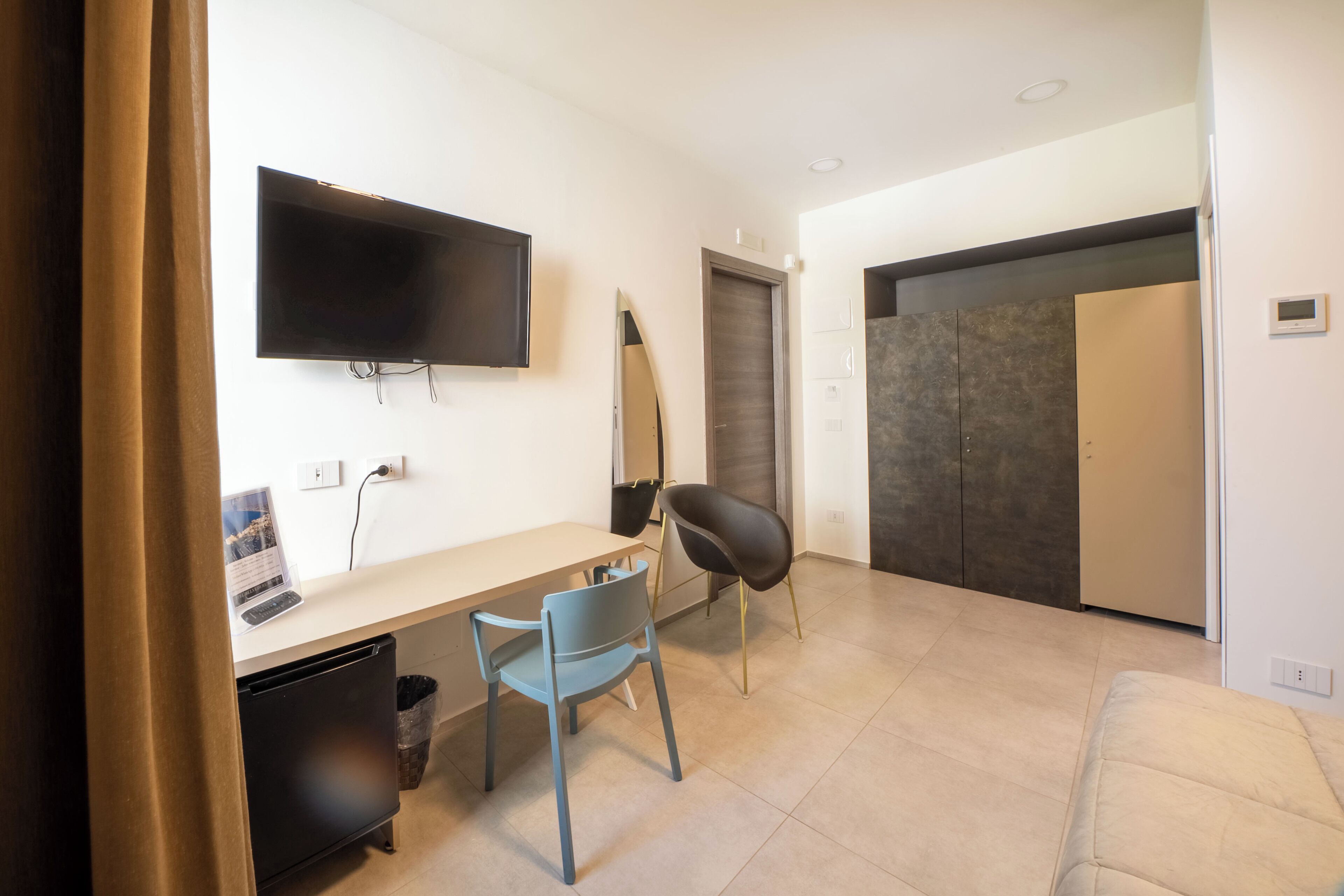 Una habitación moderna con televisión de pantalla plana, un escritorio con silla, un refrigerador pequeño y una puerta que da a otra habitación.