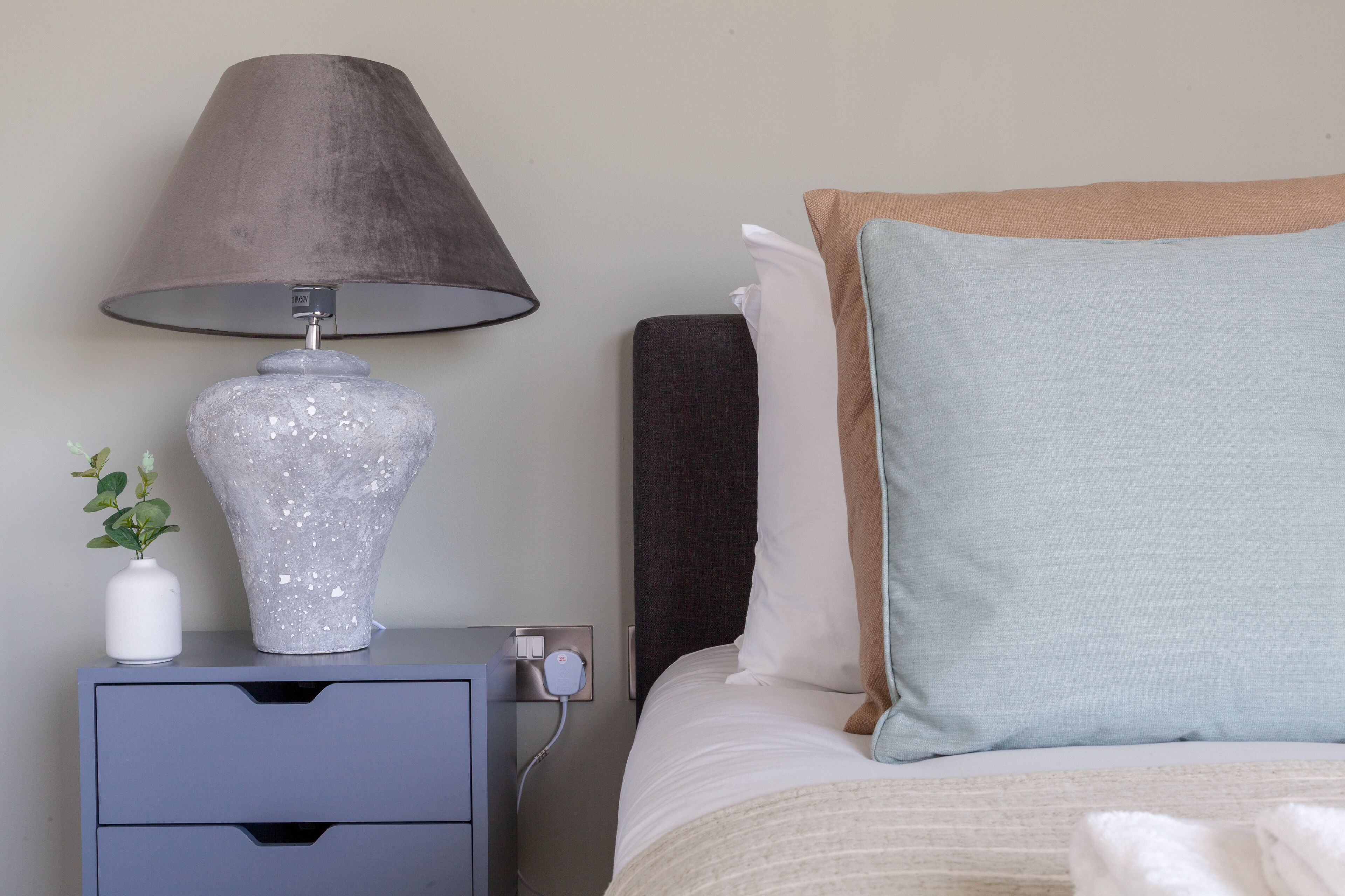 A neatly arranged bedroom with a bed, a bedside table with a lamp, a vase with a plant, and a power outlet.