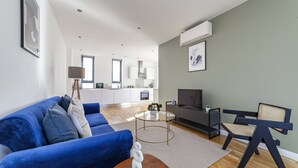 Ett modernt vardagsrum med en blå soffa, ett soffbord i glas och en unik stol i svart och beige. I bakgrunden syns ett kök med vita skåp och ett stort fönster.