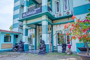 A building with a sign reading "Cathaleya" and a security sign. There are two statues in front, a motorcycle parked to the left, and potted plants with flowers.