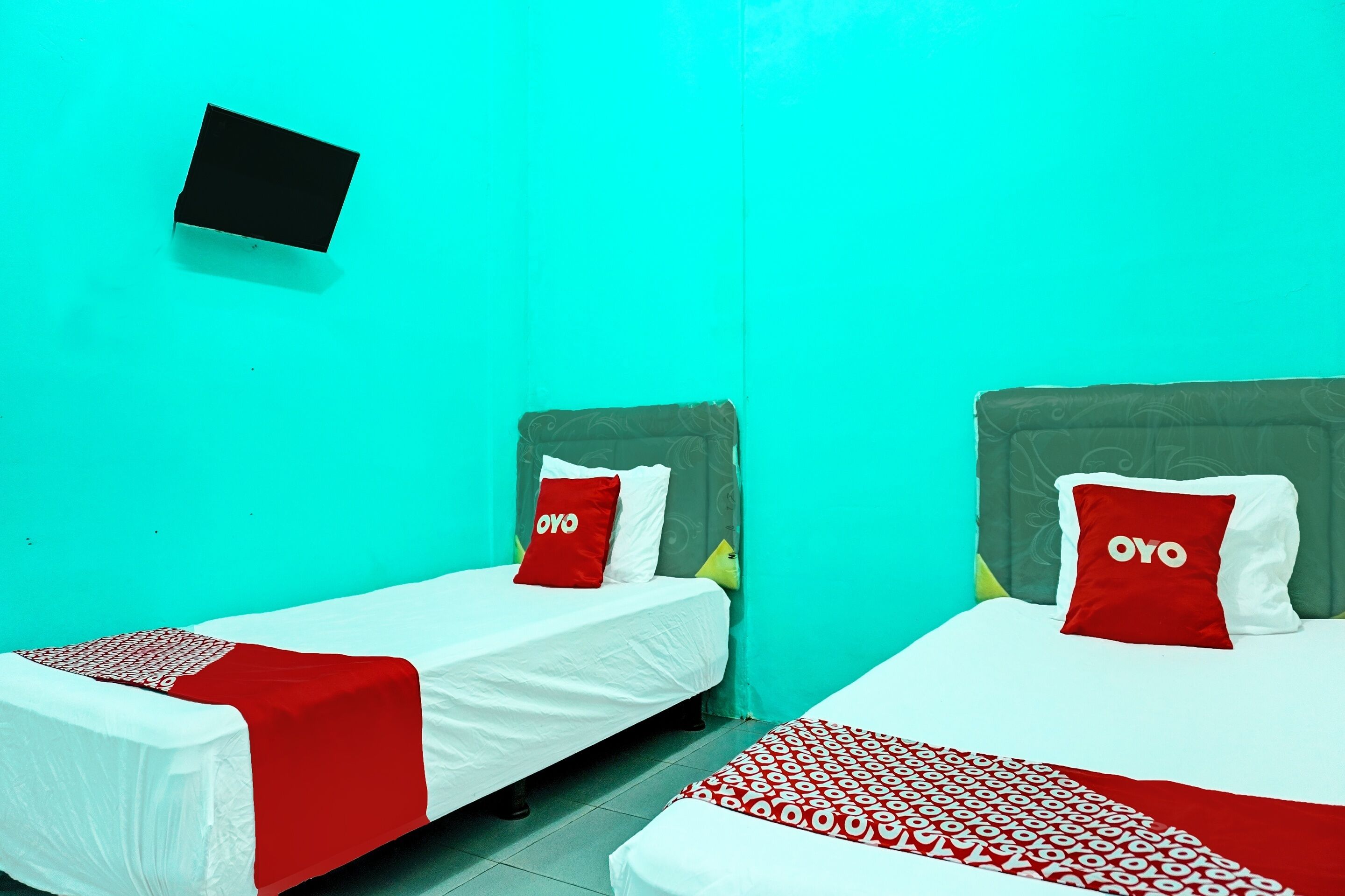Deux lits simples avec une literie rouge et blanche, un téléviseur fixé au mur et un mur de couleur turquoise.