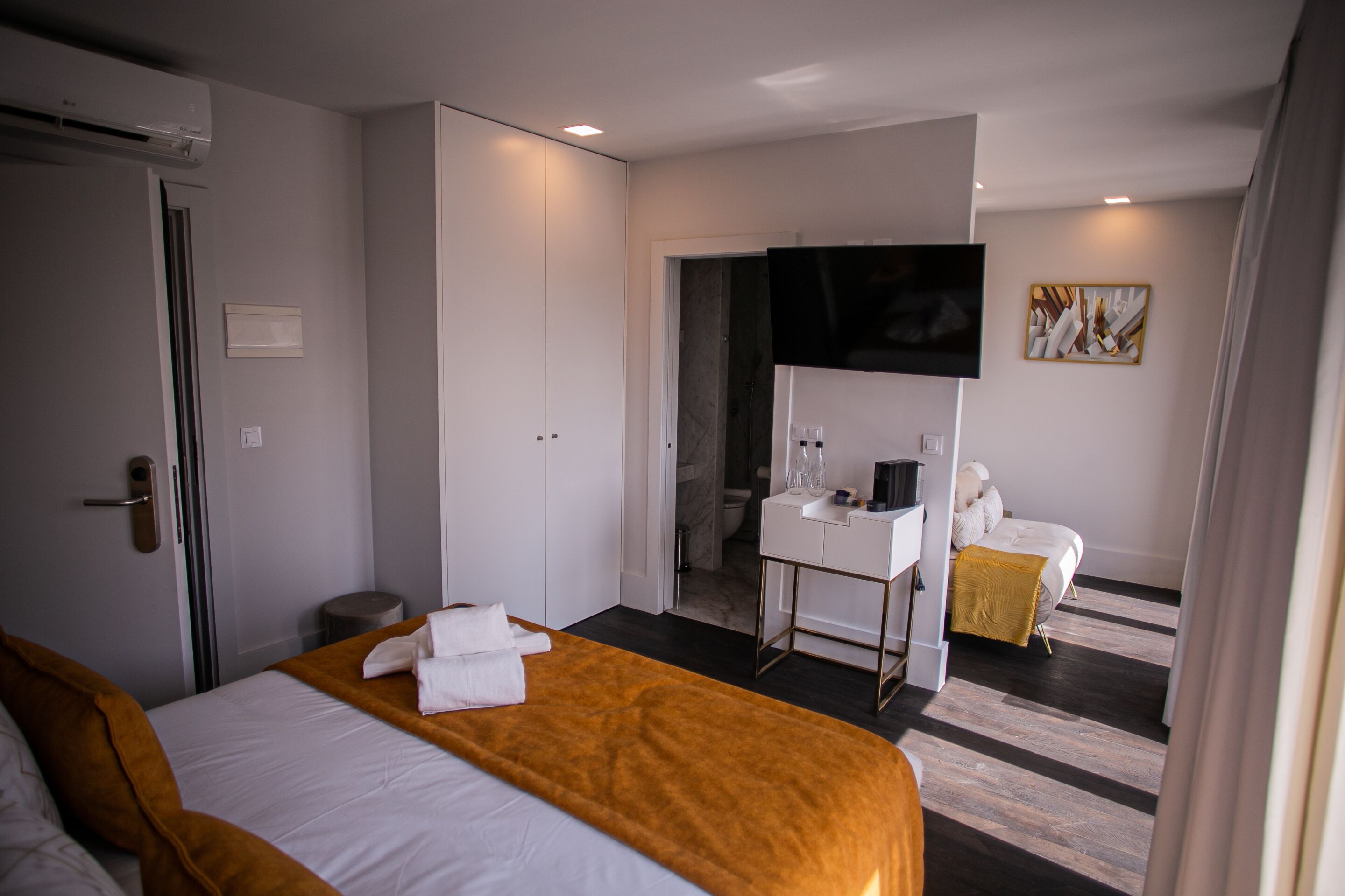 A hotel room with a large bed, a bedside table with a mirror, a small desk, a TV mounted on the wall, and a bathroom visible through an open door.