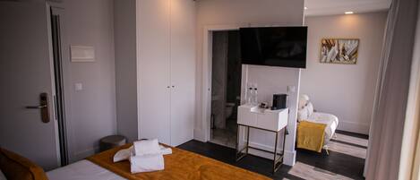 A hotel room with a large bed, a bedside table with a mirror, a small desk, a TV mounted on the wall, and a bathroom visible through an open door.