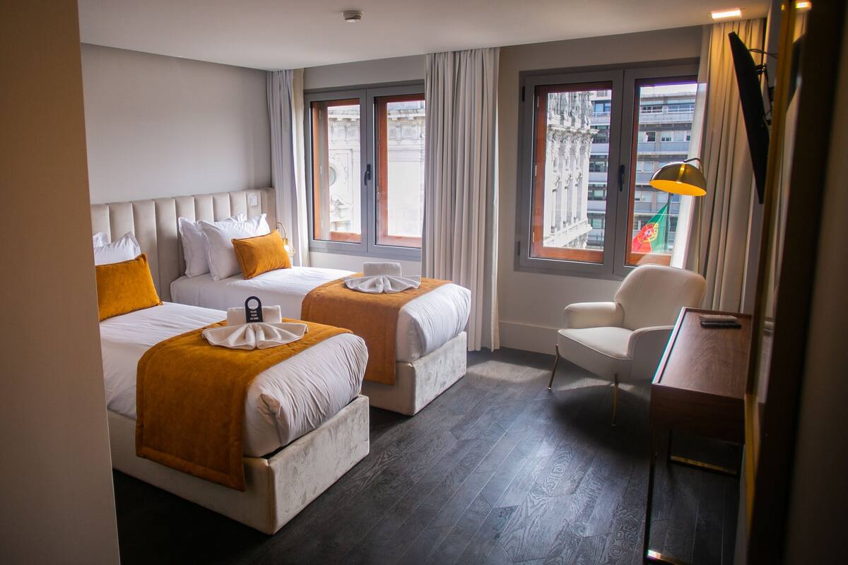 Une chambre d’hôtel avec deux lits, une chaise, un bureau et de grandes fenêtres avec des rideaux.