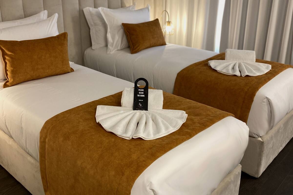 Une chambre d’hôtel avec deux lits, chacun doté d’une tête de lit marron et de draps blancs. Sur l’un des lits, on peut lire une pancarte indiquant : « Veuillez faire le ménage dans la chambre. »