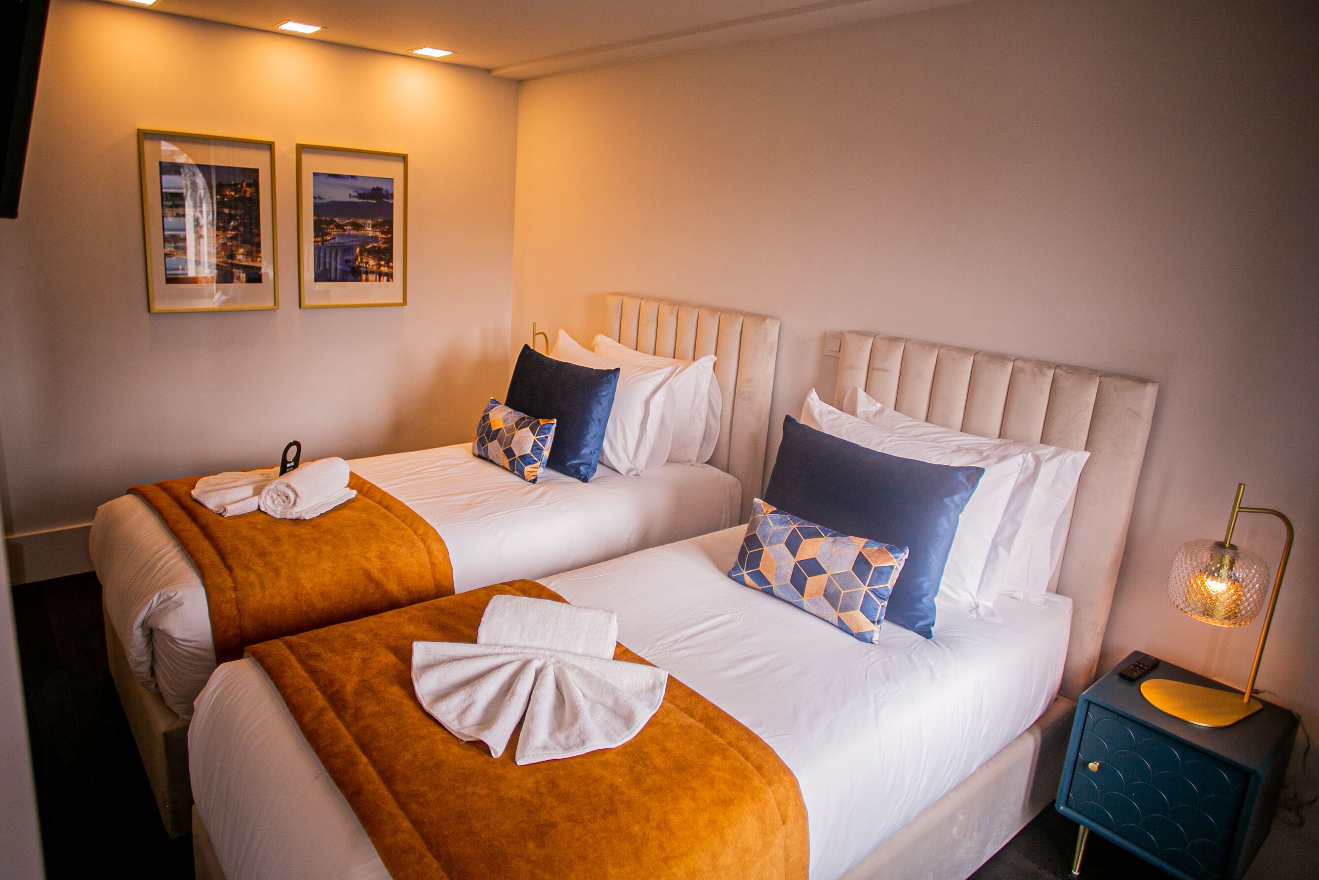 Ein Hotelzimmer mit zwei Betten, jeweils mit weißen Bettwäsche und einer braunen Tagesdecke. An der Wand befinden sich gerahmte Bilder, ein Nachttisch mit Lampe und ein blauer Schrank.
