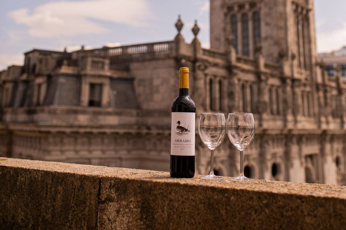Une bouteille de vin et deux verres posés sur une corniche en pierre, avec un bâtiment historique en arrière-plan.