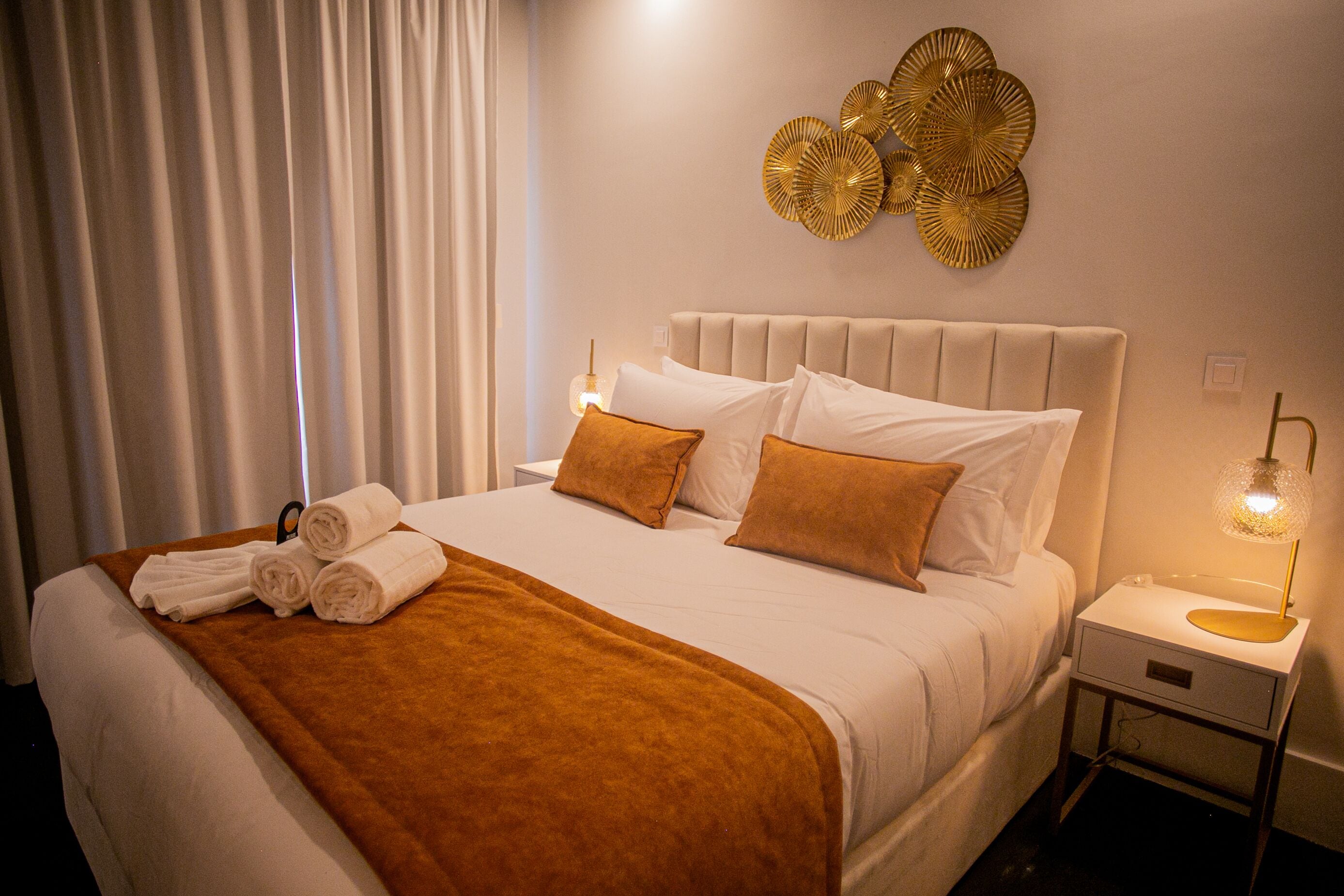 Una cama bien hecha con una manta marrón, almohadas blancas y un cabecero. Se observa una mesita de noche con una lámpara y una pieza decorativa en la pared superior.