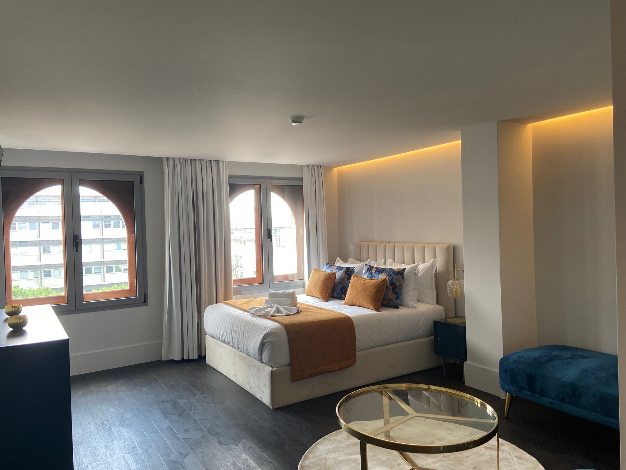 Une chambre d’hôtel moderne dotée d’un grand lit, d’une table basse en verre et offrant une vue sur la ville grâce à de grandes fenêtres en arc de cercle.