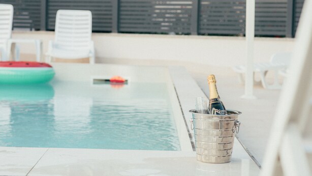 Una zona de piscina con una botella de champán en un cubo de metal, sillas reclinables blancas y un flotador verde.