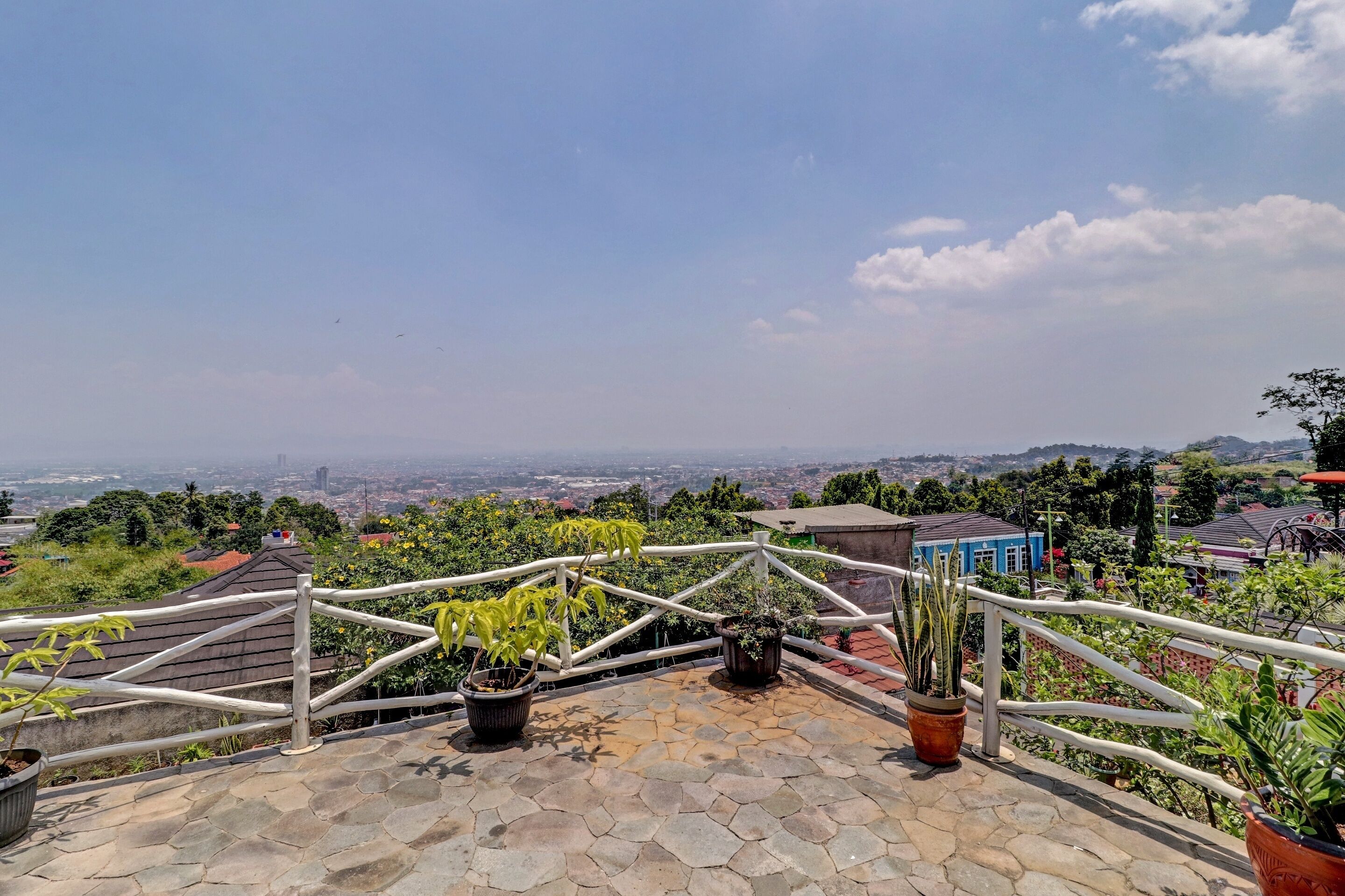 A rooftop terrace with potted plants, a white railing, and a view of a cityscape.