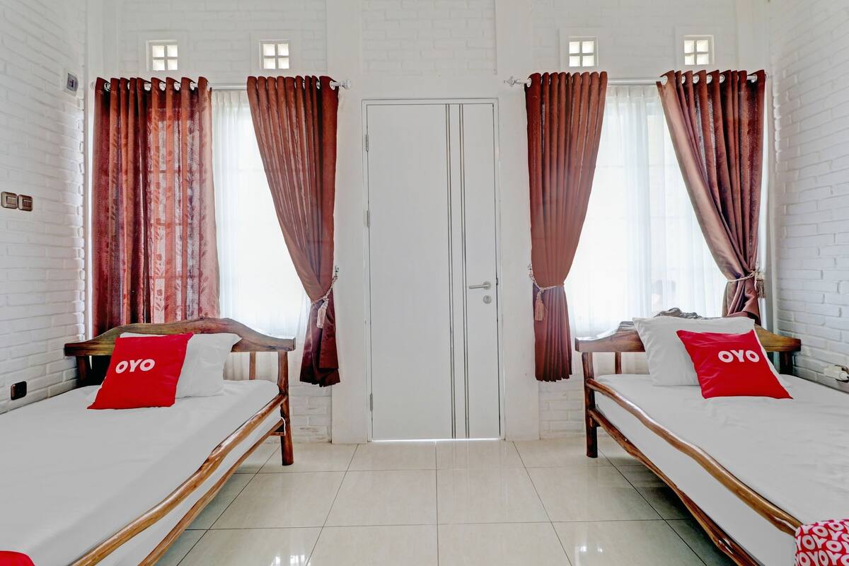 A hotel room with two beds, each with a wooden frame and white bedding. There are red pillows with the word "OYO" on them. The room has white brick walls, a white door, and curtains with a floral pattern.