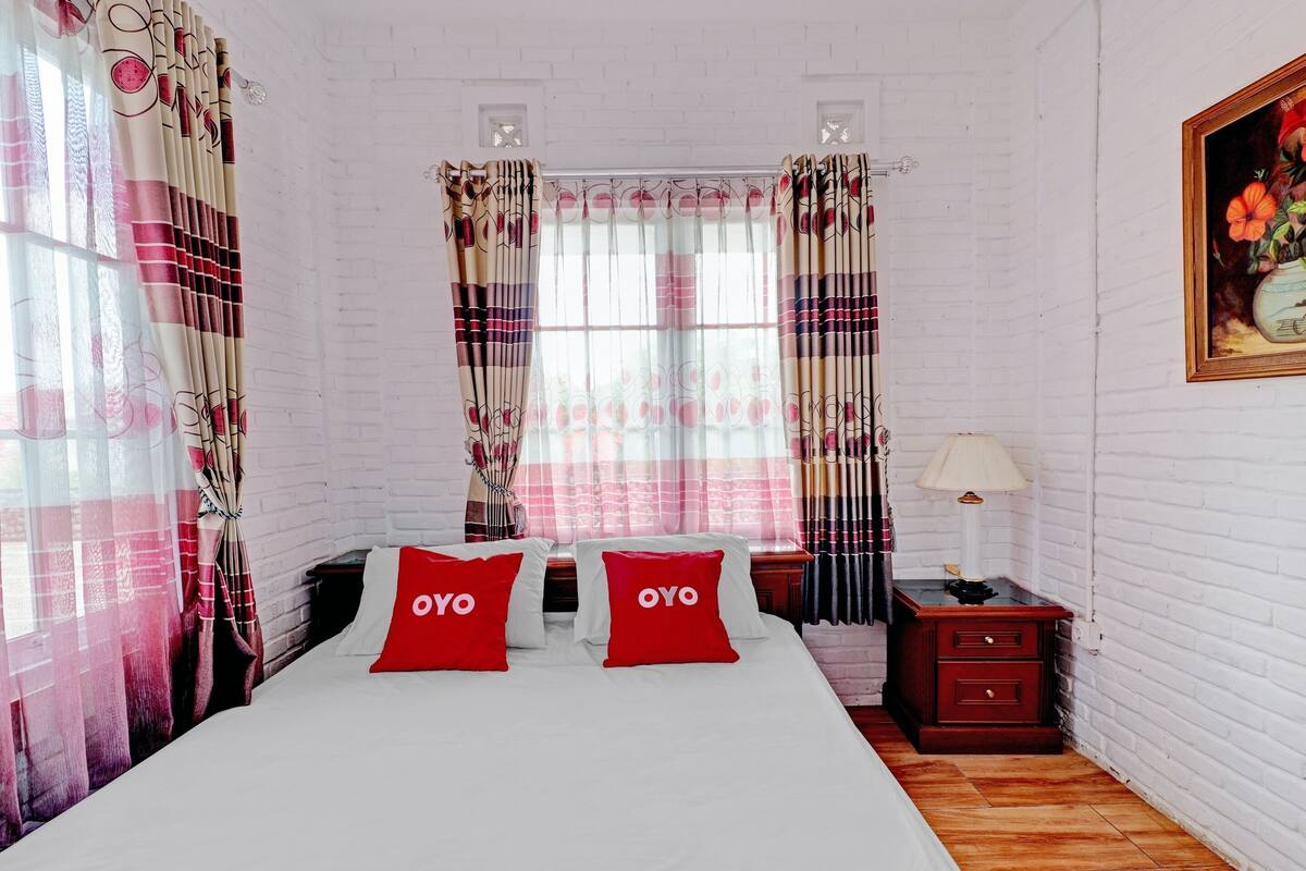 A hotel room with a bed, two red pillows with "OYO" text, a nightstand with a lamp, and a painting on the wall.