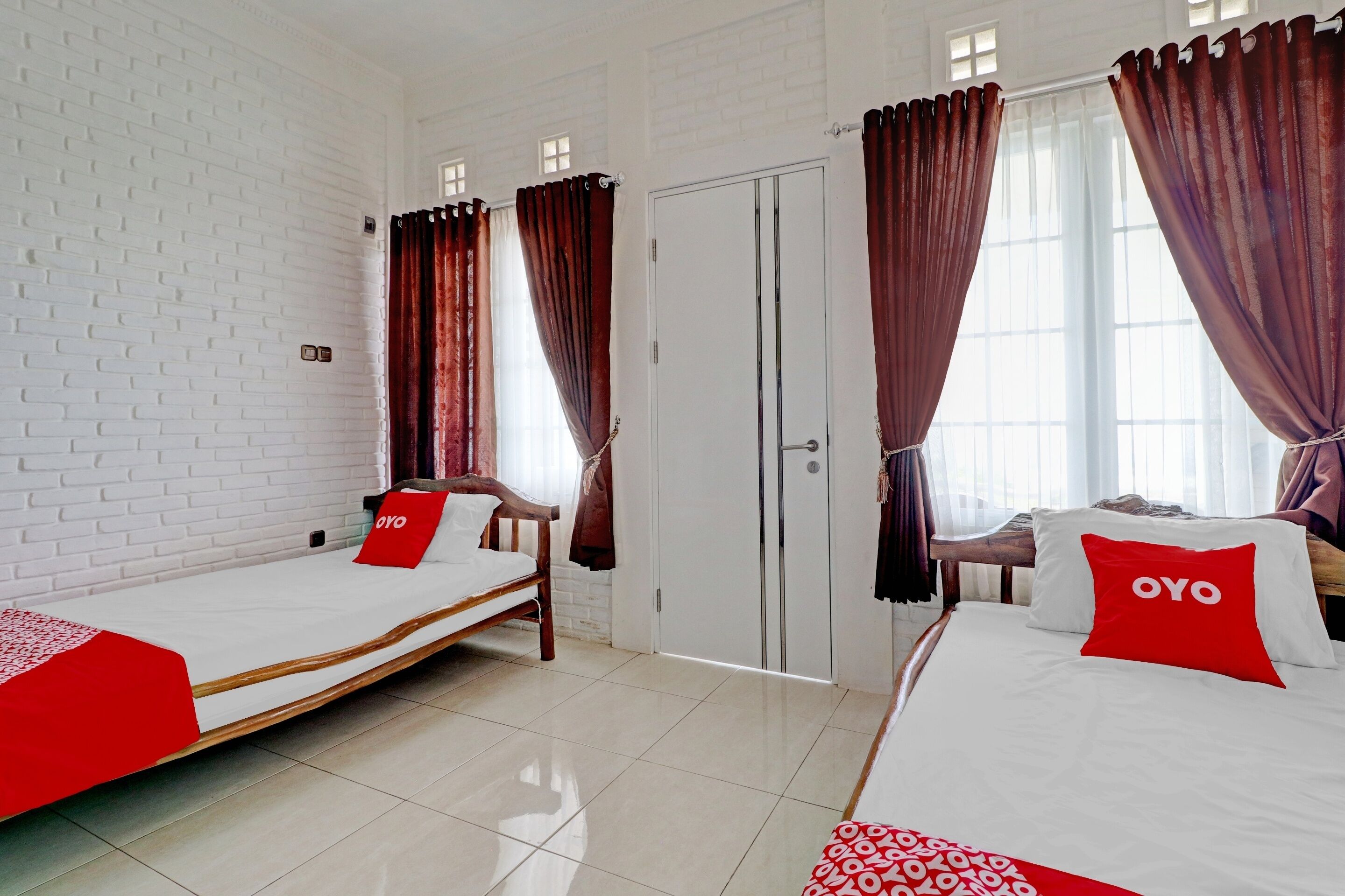 Une chambre d’hôtel avec deux lits simples, chacun agrémenté d’un oreiller rouge à l’effigie de la marque OYO, de draps blancs et d’une couverture à motifs rouges et blancs. La chambre a des murs en briques blanches, une porte blanche et de grandes fenêtres avec des rideaux bruns.