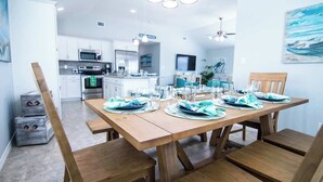 A dining area with a wooden table set for four, surrounded by wooden chairs. A kitchen is visible in the background with white cabinets and stainless steel appliances. - Off da Hook Private Heated Pool Pet Friendly (Port Aransas)