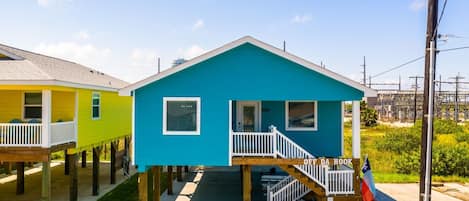 A two-story house with a blue exterior and a white porch, elevated on stilts with a wooden deck.